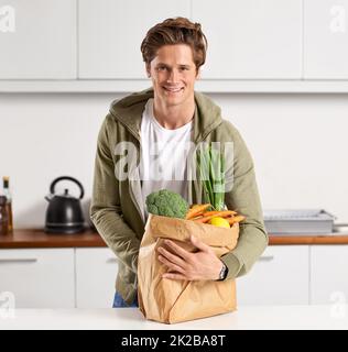 Je suis fier de ce qui va dans mon corps. Un jeune homme dans une cuisine tenant un sac de papier brun rempli de légumes. Banque D'Images