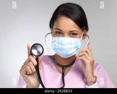 Vous avez un cœur fort. Portrait rogné d'une jeune travailleuse de santé attrayante portant un masque et tenant un stéthoscope en studio sur fond gris. Banque D'Images