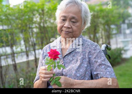 Femme asiatique âgée ou âgée, portant une rose rouge sur un fauteuil roulant dans le parc. Banque D'Images