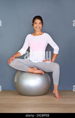 Le corps réalise ce que l'esprit croit. Prise de vue en longueur d'une jeune femme sportive assise sur un ballon pilates sur un fond gris. Banque D'Images