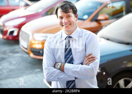 Il peut vous donner une bonne affaire de voiture. Un homme souriant avec assurance en se tenant dans le lot de sa concession. Banque D'Images