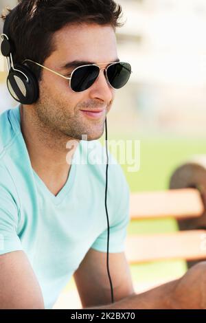 Écoutez ses morceaux préférés. Un jeune homme souriant portant des lunettes de soleil assis sur un banc dans un parc et écoutant de la musique à travers ses écouteurs. Banque D'Images