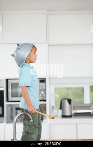 Je suis un chevalier - imagination. Un jeune garçon mignon utilisant une passoire de riz comme un casque et une cuillère en bois comme une épée dans la cuisine. Banque D'Images