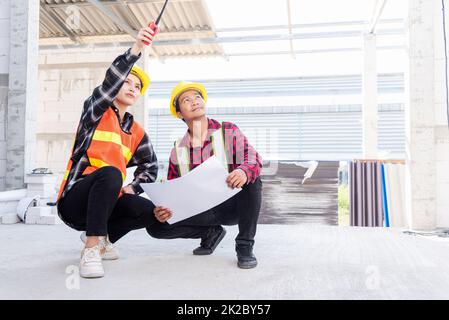 L'architecte et le client discutent de l'aide à la création d'un plan avec le plan du bâtiment sur le chantier de construction. Un homme et une femme de travail de contremaître d'ingénieur asiatique se rencontrent pour discuter du projet de papier à dessin, travail d'équipe Banque D'Images