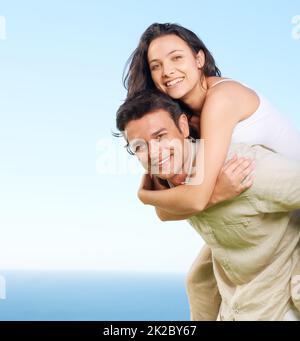 La prendre pour voir la vue. Un jeune homme qui fait un coggysoutenant sa petite amie avec une vue sur l'océan en arrière-plan. Banque D'Images
