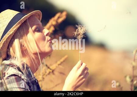 Elle se soucie de ses soucis Belle jeune femme portant une fedora de paille et soufflant à un pissenlit. Banque D'Images