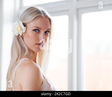 Beauté et pureté. Portrait d'une femme attrayante avec une rose blanche dans ses cheveux tout en se tenant dans sa chambre à coucher. Banque D'Images