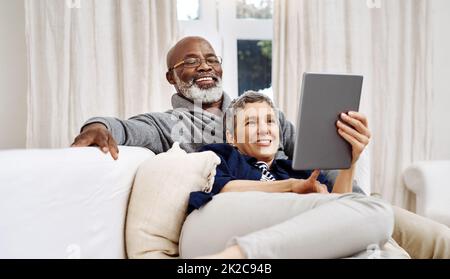 Passer la journée ensemble à la maison. Prise de vue d'un couple aîné affectueux utilisant une tablette tout en se relaxant sur le canapé à la maison. Banque D'Images