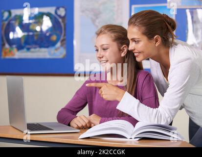 Stimuler les jeunes esprits. Une belle jeune enseignante penchée sur son élève et pointant vers quelque chose sur son écran d'ordinateur portable. Banque D'Images