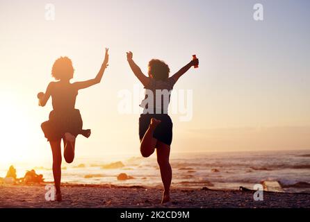 Aimer la vie nous vivons ensemble comme des amis. Vue arrière de deux jeunes femmes méconnues qui sautent dans la joie tout en marchant le long de la plage au coucher du soleil. Banque D'Images