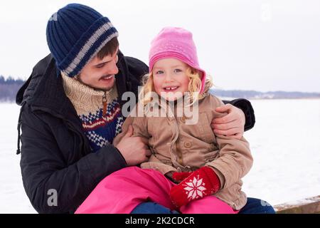 J'adore passer du temps avec mon père. Une petite fille qui s'assoit avec joie sur ses pères se lap dehors lors d'une journée d'hivers croustillante. Banque D'Images