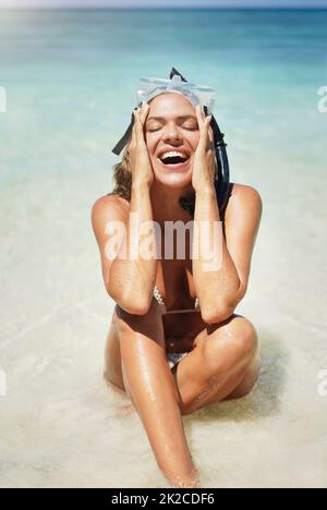 C'est toujours amusant au soleil. Photo courte d'une jeune femme attirante en équipement de plongée qui rit tout en étant assise sur la plage. Banque D'Images