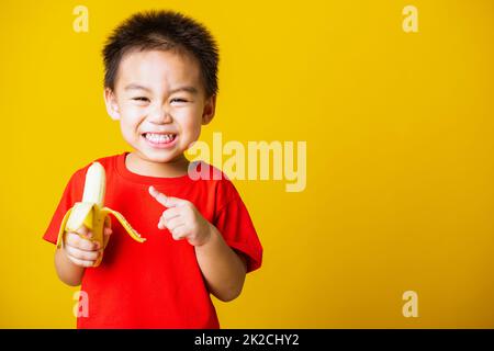 enfant mignon petit garçon sourire attrayant portant t-shirt rouge jouer contient la banane pelée pour manger Banque D'Images