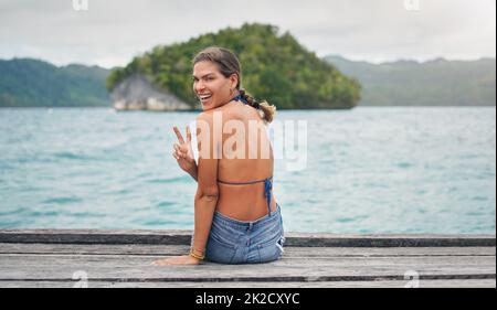 Vivre ma meilleure vie. Portrait court d'une jeune femme attrayante assise sur le bord d'une promenade et faisant un geste de signe de paix. Banque D'Images