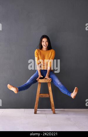 Feel like a kid again. An attractive young woman sitting playfully on a stool against a gray wall. Banque D'Images