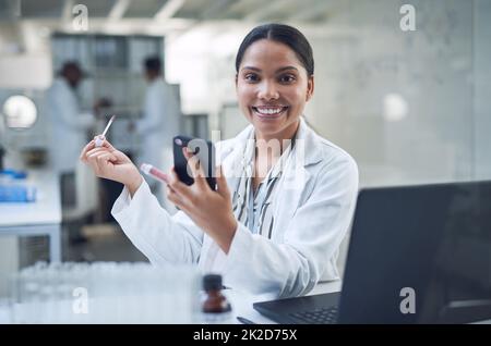 Nos cosmétiques sans cruauté vous permettent de garder l'air bien moins la culpabilité. Photo d'un jeune scientifique appliquant du maquillage à l'aide d'un smartphone tout en effectuant des recherches dans un laboratoire. Banque D'Images