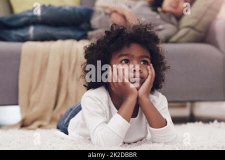 Je me demande si therell être une partie deux. Photo d'un adorable petit garçon allongé sur un tapis et regardant la télévision à la maison. Banque D'Images