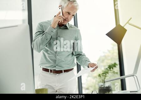 En parlant du contrat. Photo d'un homme d'affaires mûr parlant au téléphone tout en travaillant debout à son bureau. Banque D'Images