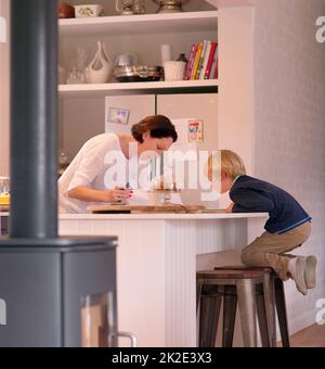 Watcha faisant, maman. Photo d'un petit garçon mignon regardant sa mère travailler dans leur cuisine à la maison. Banque D'Images