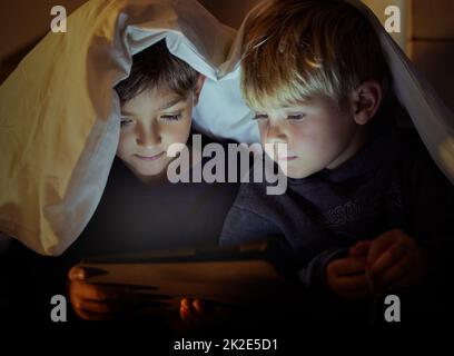 Les yeux des enfants voient tout frais et plein d'émerveillement. Photo de deux adorables petits garçons utilisant une tablette numérique sous une couverture fort la nuit dans leur chambre. Banque D'Images