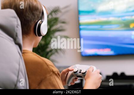 Vue arrière d'un jeune garçon avec casque et joystick jouant à la console de jeux vidéo. Banque D'Images