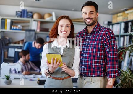 Étaient un duo de design dynamique. Portrait de deux jeunes créateurs de mode debout dans l'atelier pendant que leurs collègues travaillent en arrière-plan. Banque D'Images