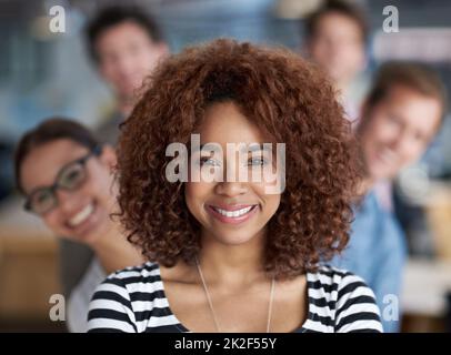 Prendre les devants en toute confiance. Jeune femme souriante regardant la caméra avec le personnel en arrière-plan. Banque D'Images