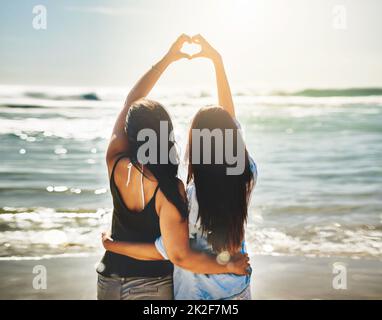 Nous partageons un amour pour les belles choses. Vue arrière de deux amis formant un coeur ensemble sur la plage. Banque D'Images