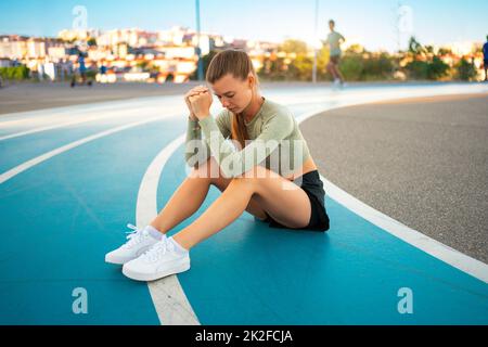 Athlète féminine fatiguée et bouleversée assise sur une piste de course à pied après des exercices de jogging. Résultat sportif déçu. Sprinter amateur sportswoman épuisé après des entraînements difficiles assis sur la piste Banque D'Images