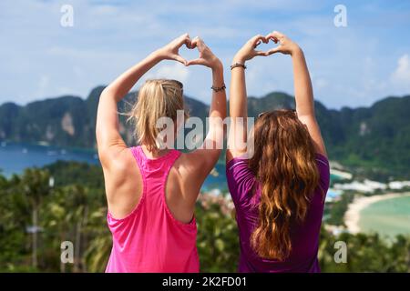 Vivez les moments qui rendent la vie magnifique. Vue arrière de deux jeunes femmes faisant un geste de coeur en faisant face à un paysage pittoresque. Banque D'Images