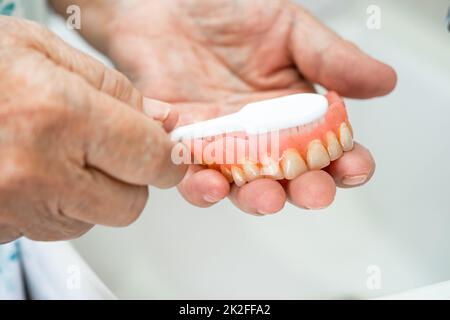 Une patiente asiatique âgée ou âgée utilise une brosse à dents pour nettoyer une prothèse partielle des dents de rechange. Banque D'Images