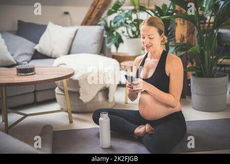 Jeune heureuse et gaie belle femme enceinte qui se fait plaisir avec sa famille et ses amis sur son téléphone portable tout en restant en forme, sportive et active pendant son congé de maternité. Grossesse, concept de yoga Banque D'Images