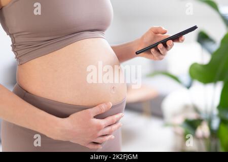 Photo en gros plan du ventre de la femme enceinte. Femme tenant et utilisant l'application de smartphone mobile à l'intérieur de la maison. Grossesse, technologie, achats en ligne, préparation et concept d'attente. Banque D'Images