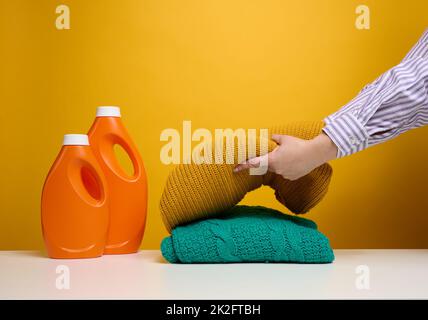 vêtements pliés lavés et grandes bouteilles en plastique orange avec support de détergent liquide sur une table blanche, fond jaune.Devoirs de routine Banque D'Images