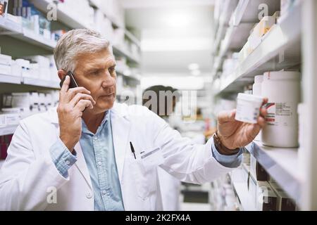 Nous avons besoin de plus de analgésiques. Photo d'un pharmacien de sexe masculin mature et concentré qui parle sur son téléphone portable tout en lisant le dos d'une boîte de médicaments. Banque D'Images