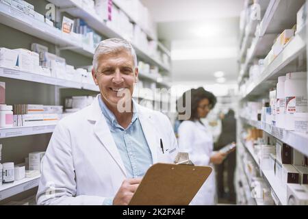 Tout en une journée de travail en tant que pharmacien. Portrait d'un pharmacien homme mature et gai tenant une planchette à pince et faisant le stock tout en regardant la caméra dans une pharmacie. Banque D'Images