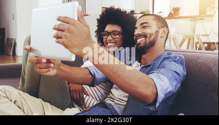 Nous prenons la journée ensemble. Prise de vue d'un jeune couple joyeux assis sur un canapé tout en prenant un autoportrait avec une tablette numérique dans le salon à la maison. Banque D'Images