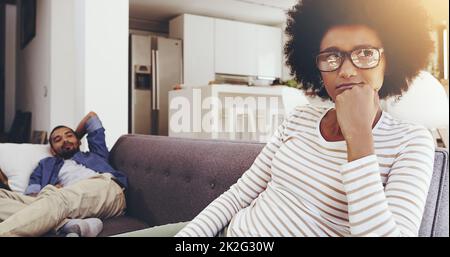 Il peut m'ennuyer tellement parfois. Prise de vue d'une femme qui a l'air agacée assise sur un canapé, avec son menton sur son poing, tout en ignorant son petit ami à la maison. Banque D'Images