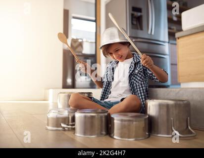 HES est un percussionniste né. Photo d'un petit garçon heureux jouant des tambours avec des pots sur le sol de la cuisine tout en portant un bol sur sa tête. Banque D'Images