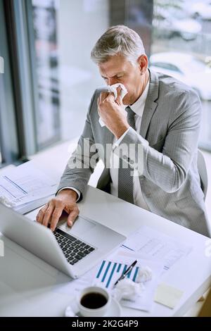 Il n'est pas productif d'être malade au travail. Photo courte d'un homme d'affaires malade qui soufflait le nez au bureau tout en travaillant sur un ordinateur portable. Banque D'Images