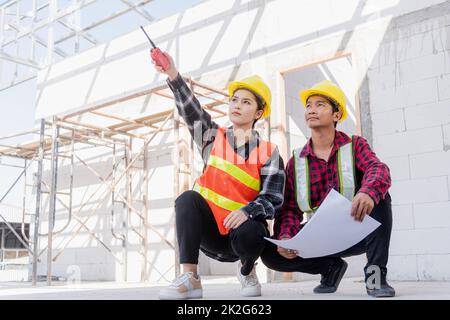 L'architecte et le client discutent de l'aide à la création d'un plan avec le plan du bâtiment sur le chantier de construction. Un homme et une femme de travail de contremaître d'ingénieur asiatique se rencontrent pour discuter du projet de papier à dessin, travail d'équipe Banque D'Images