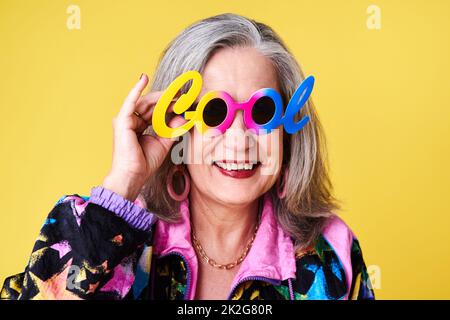 Je joue toujours au frais. Portrait d'une femme âgée confiante et élégante portant des lunettes de soleil se posant sur un fond jaune. Banque D'Images