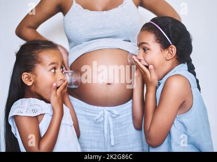 Mouvement theres dans là. Photo de deux petites filles gaies debout à côté de leur mère tout en mettant un verre sur son ventre enceinte pour écouter le mouvement à la maison. Banque D'Images