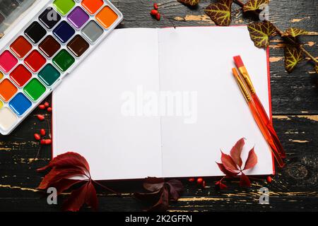 Opened notebook, watercolor paints and brushes on dark wooden background Stock Photo