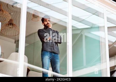 Réfléchir à sa prochaine grande réalisation. Photo d'un jeune homme d'affaires qui a l'air réfléchi tout en se tenant debout sur un mur de verre dans un bureau. Banque D'Images