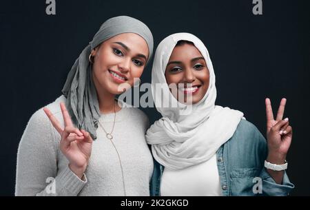 Tendances du millénaire. Coupe courte de deux jeunes femmes séduisantes portant des hijabs et debout ensemble tout en montrant un signe de paix dans le studio. Banque D'Images