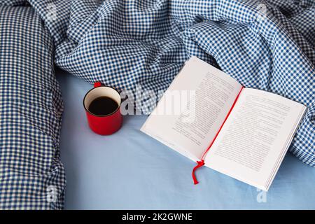 Ukraine, Rivne - 11 août 2021: Vue de dessus d'une couverture confortable, un livre ouvert et une tasse de café rouge chaud sur le lit lors d'une froide journée d'hiver. Concept détente et hygge. Vue de dessus avec espace de copie. Banque D'Images