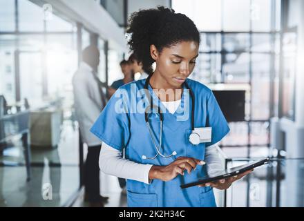 Moins de papier, plus de diagnostic. Prise de vue d'un jeune médecin utilisant une tablette numérique dans un hôpital avec ses collègues en arrière-plan. Banque D'Images