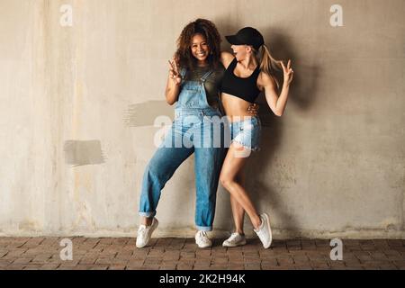 Nous savons comment Amuse-toi bien. Portrait de deux jeunes femmes gaies posant pour une photo tout en se penchant contre un mur à l'extérieur pendant la journée. Banque D'Images