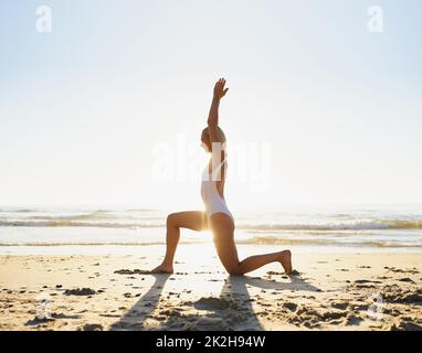 Commencer ma journée du bon pied. Prise de vue en longueur d'une jeune femme attirante faisant du yoga tôt le matin sur la plage. Banque D'Images